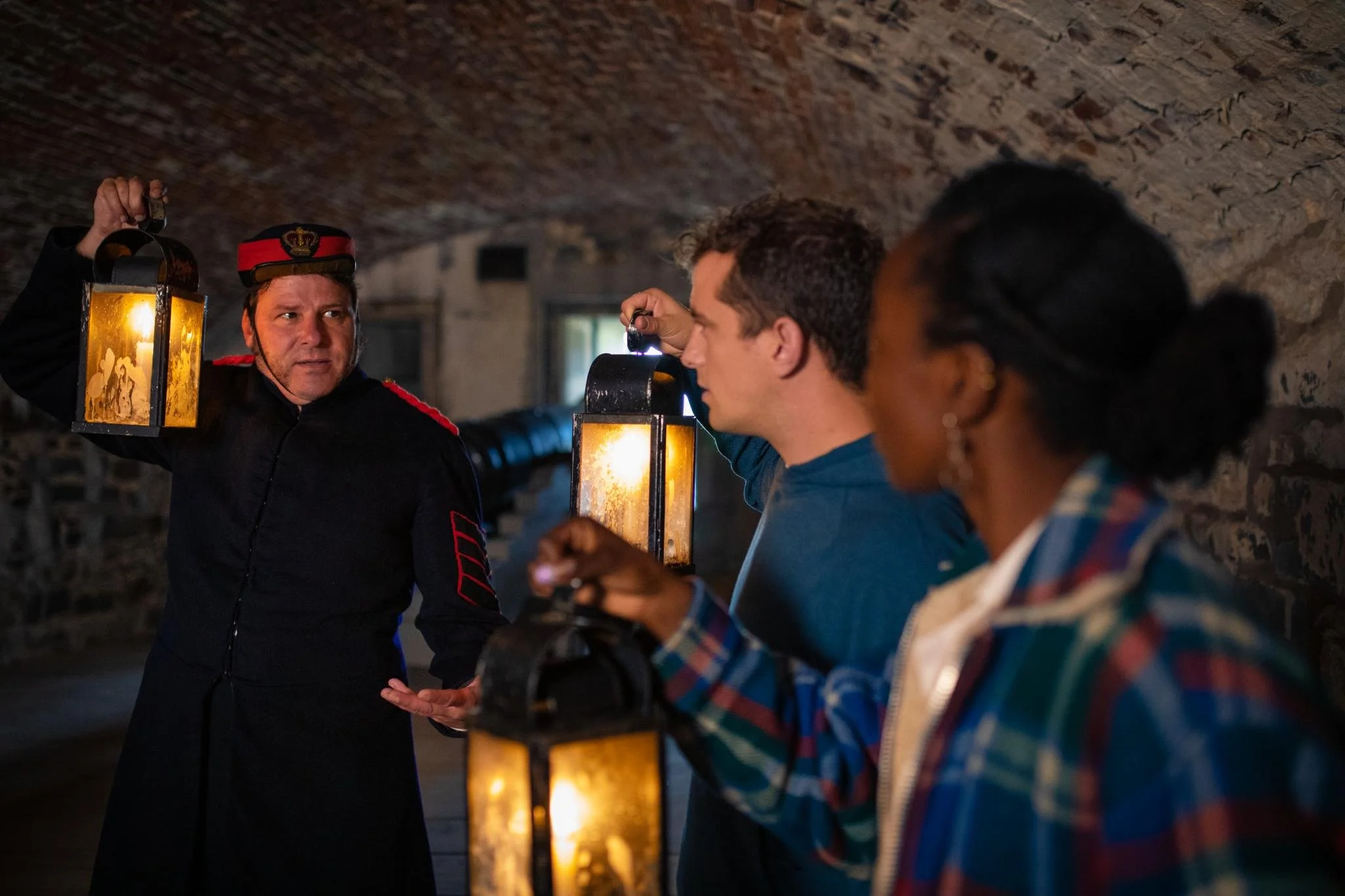 A man dressed as an officer shows a lantern to a group of people in a tunnel with a low, arched stone ceiling.