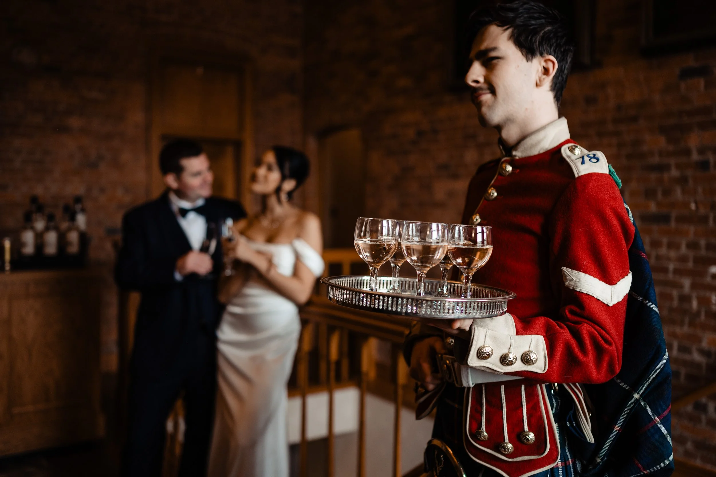 A waiter in a red uniform holding a tray of glasses at a formal event. In the background, a couple dressed in formal attire is talking and smiling.