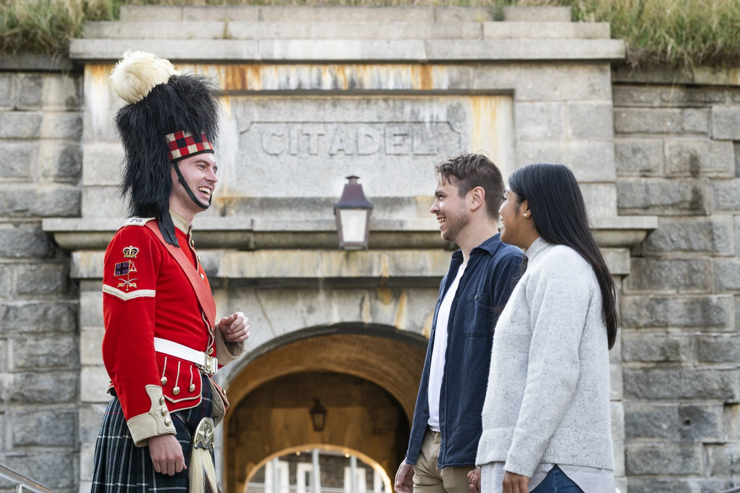 A man dressed in traditional 78th Highlander uniform, including a red jacket, tartan kilt, and a feathered hat, talking to a young man and woman in front of a stone archway with a sign that says 'CITADEL'.