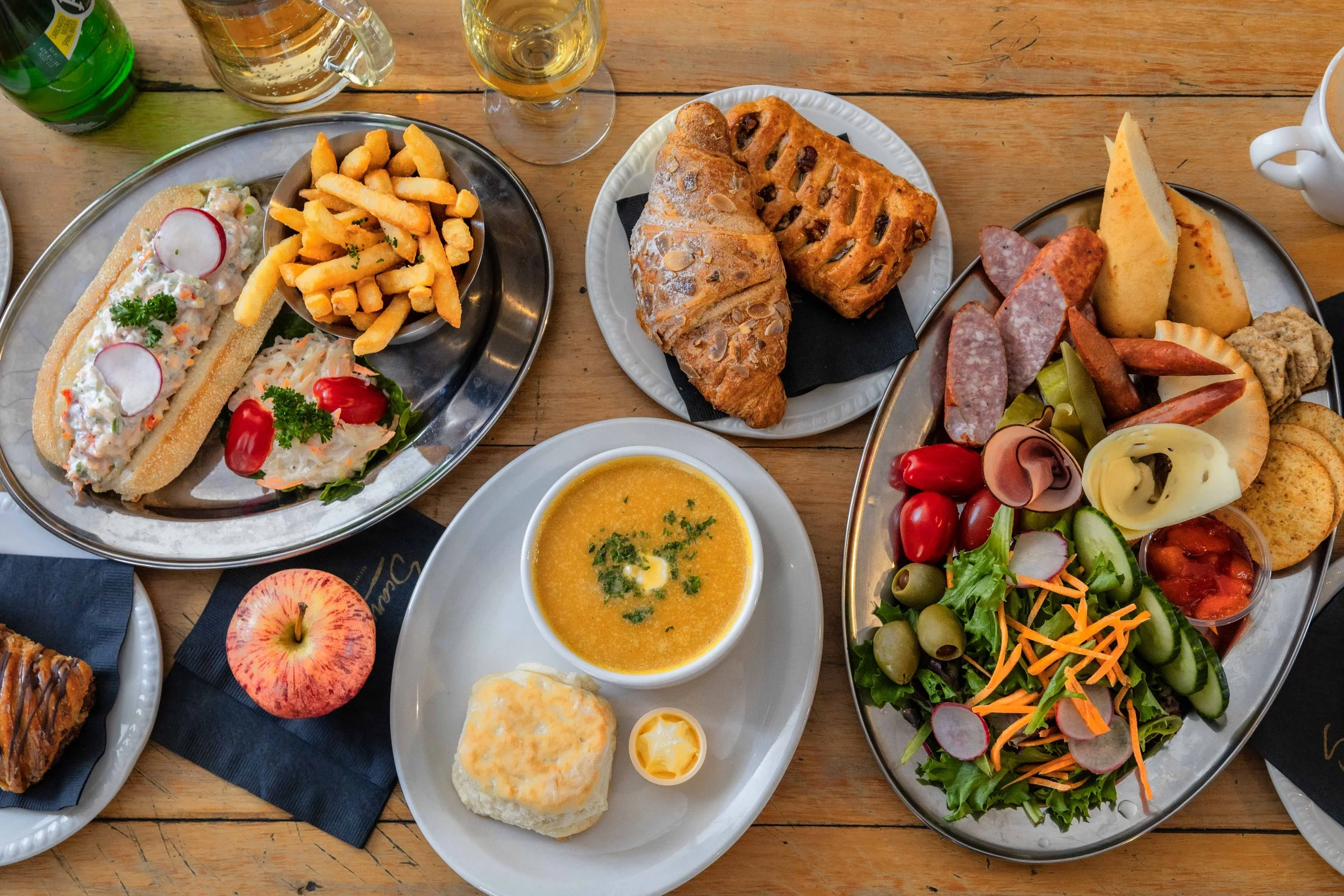 A table with various foods including a sandwich, fries, salad, bread, cheese, cold cuts, soup, and an apple, along with drinks like beer and soda.