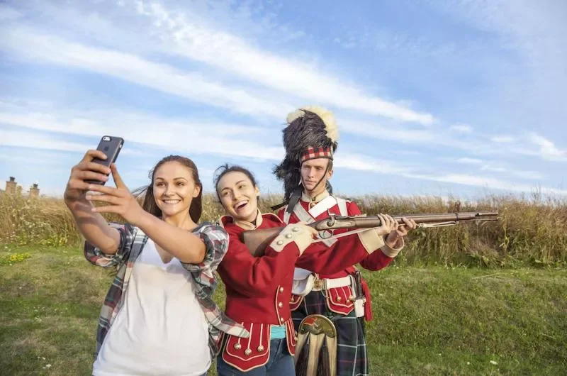 Two young women taking a selfie with a man dressed in a historical soldier costume holding a musket in an outdoor field.
