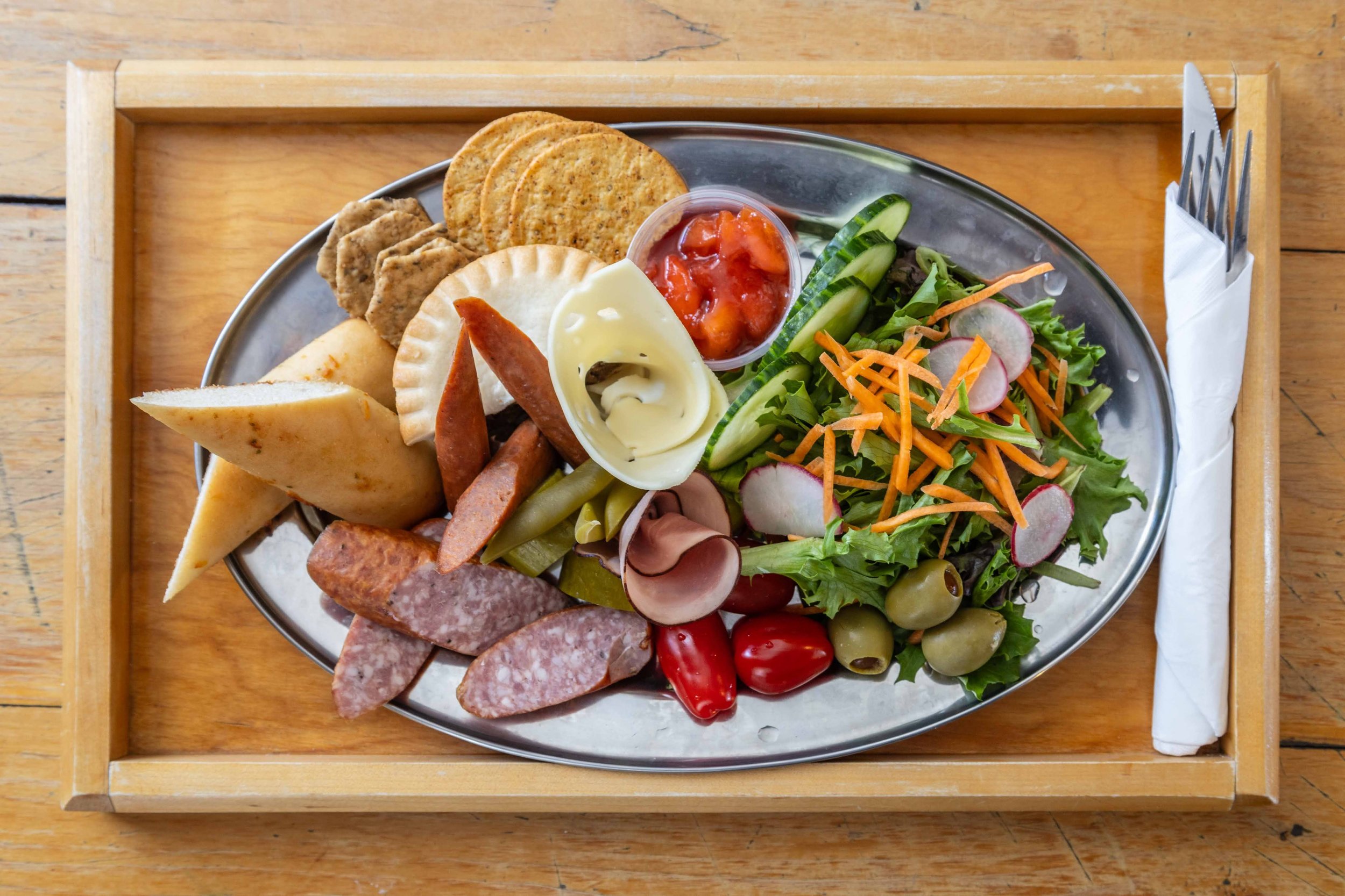 Charcuterie plate with cheese, crackers, vegetables, and assorted meats, served on a metal tray with a side of salsa, on a wooden table.
