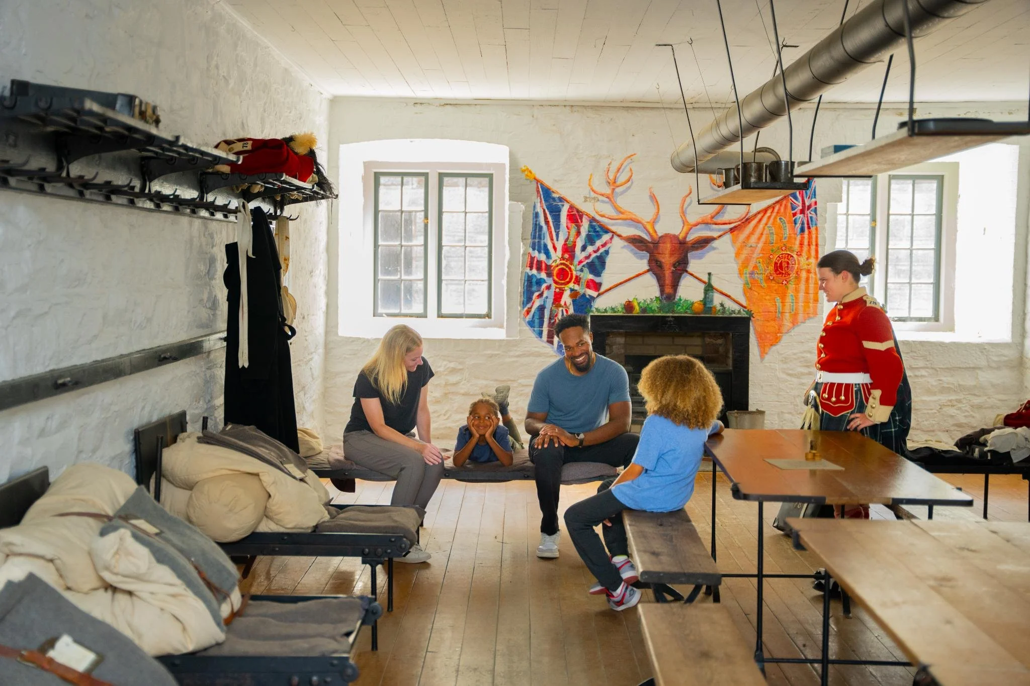 A group of people are gathered inside a room with white brick walls and large windows. Some are sitting and others standing, engaged in conversation and smiling. One person is dressed in a traditional British guard uniform. The room is decorated with flags and a large deer head mural on the wall.