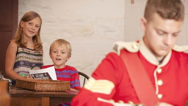 A young girl and boy looking at a man in a red uniform, who is writing or drawing, in a room with white brick walls.