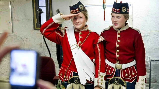Two young children dressed in historic costumes, one saluting, in a room with white brick walls.