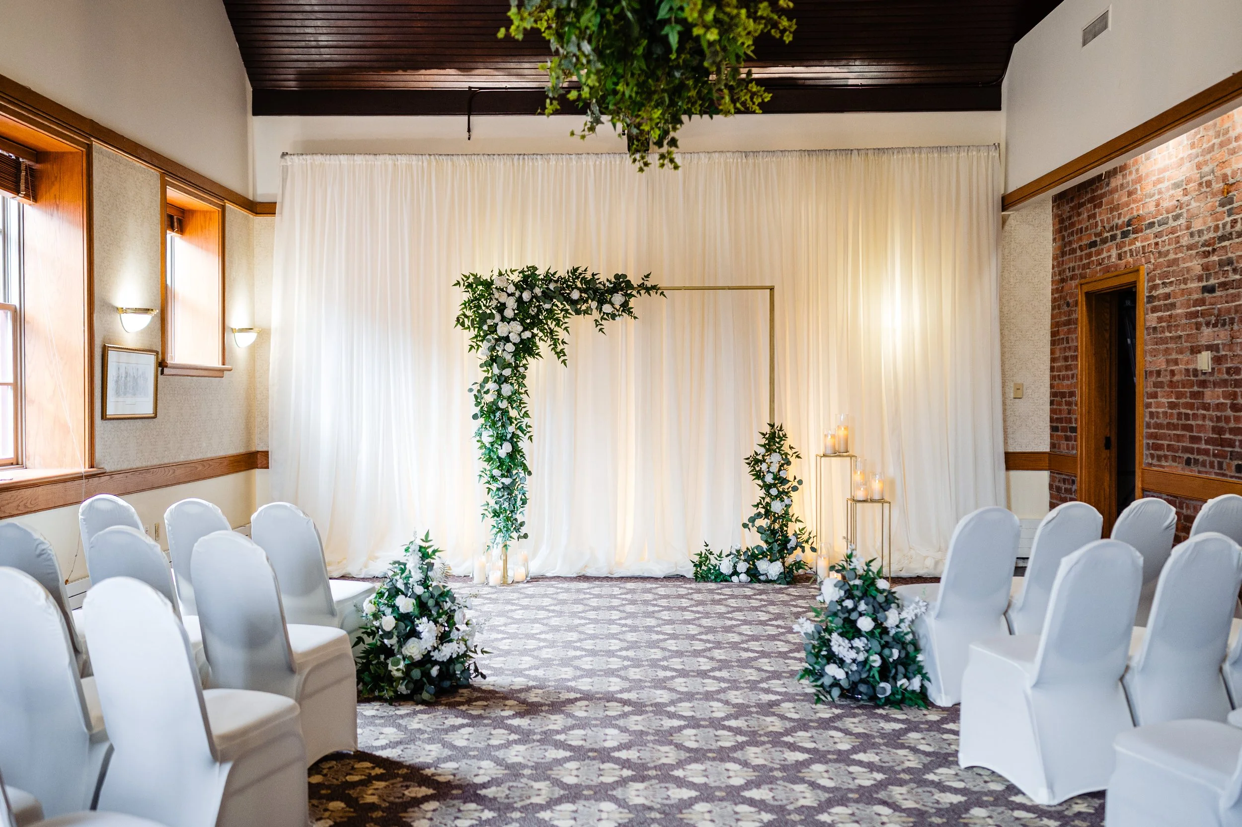 Indoor wedding ceremony setup with white chairs arranged in rows facing a decorated altar with white flowers and green foliage, draped white curtains, and candles on stands, with a brick wall and windows on the sides.
