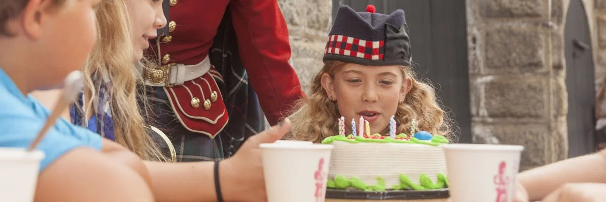 Girl in a black and red Scottish hat blowing out birthday candles on a cake surrounded by friends.