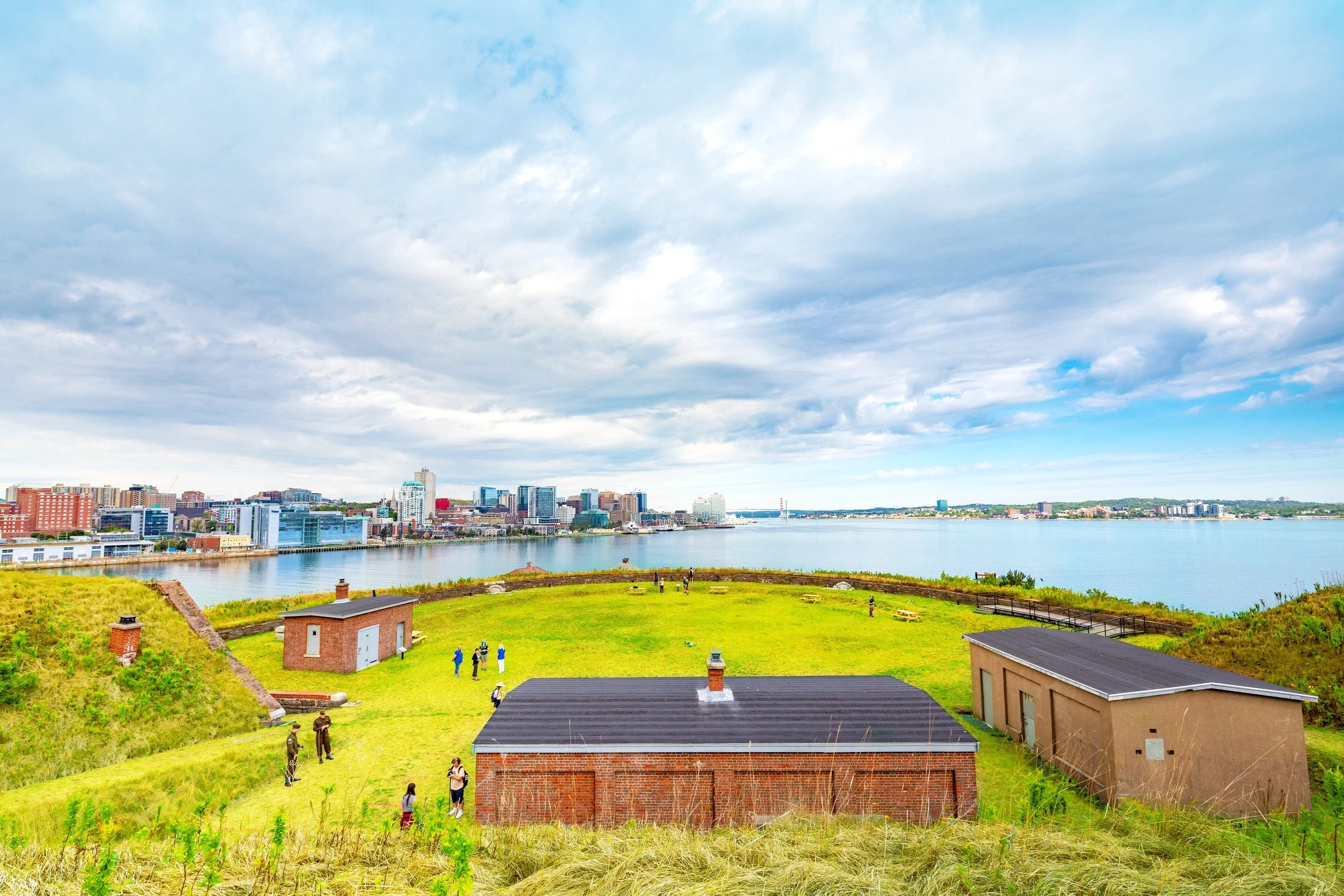 A city skyline viewed from a grassy hill with historic brick buildings in the foreground, and taller modern buildings along a river with a partly cloudy sky.