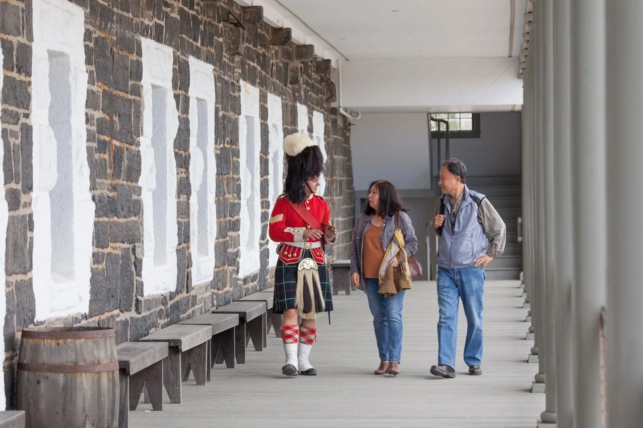 A person dressed in a 78th Highlander uniform talking with a couple walking along a corridor with wooden benches and a brick wall.
