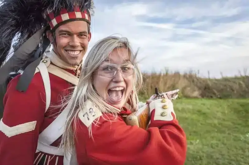 Two people in historical military costumes, smiling outdoors with a cloudy sky and grassy field in the background.