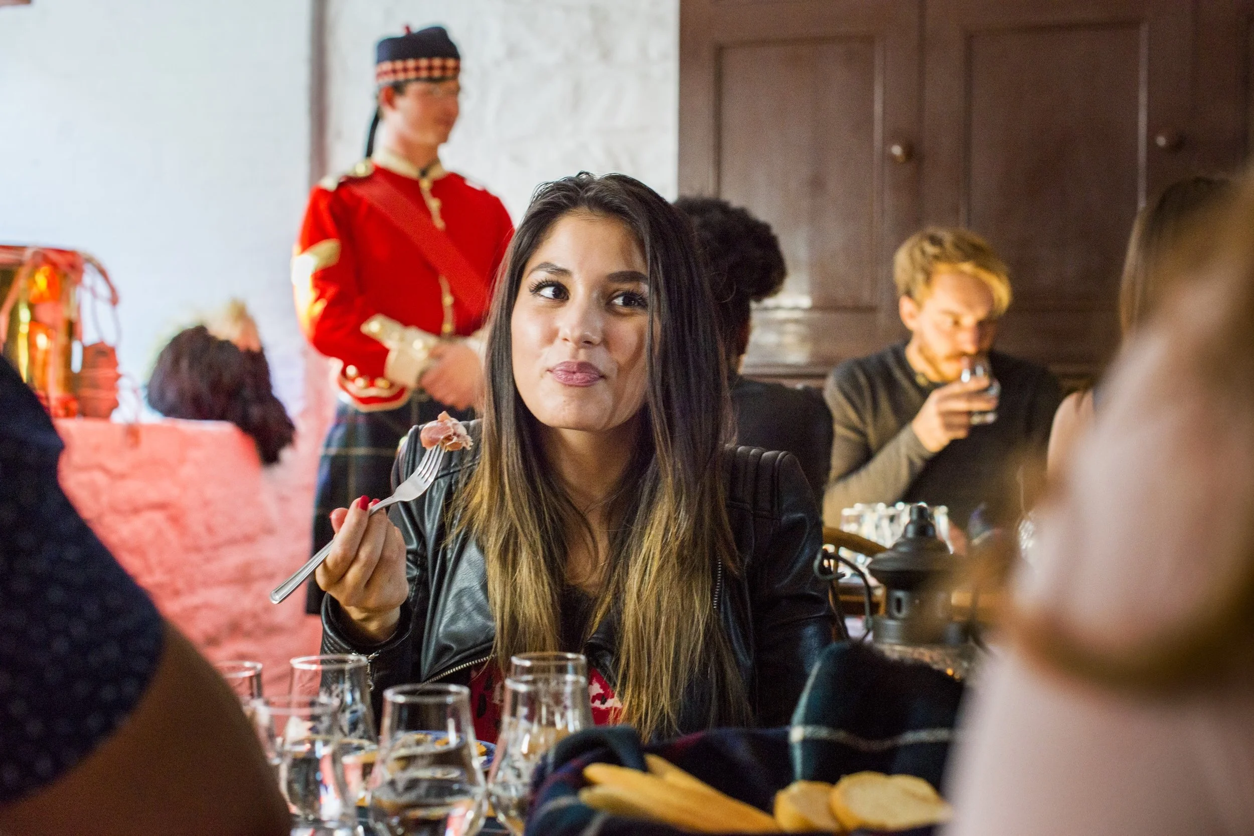 A woman with long dark hair and light brown skin sitting at a table in a restaurant, holding a fork with a piece of food. She is smiling and looking slightly to the side. Behind her, a person dressed in a red costume that resembles a soldier or royal guard is standing, holding a white napkin. Other people are sitting at the table, and there are glasses of water and plates of food, including bread rolls, on the table.