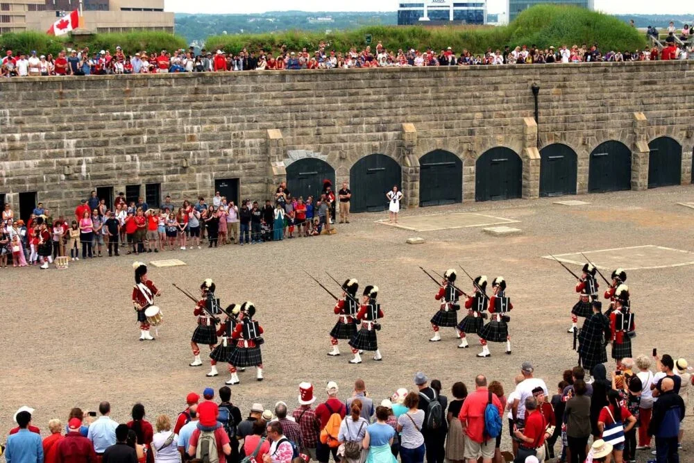 A group of dancers in traditional dress performing for an outdoor audience, with spectators gathered around and above, outside a stone fort or wall.