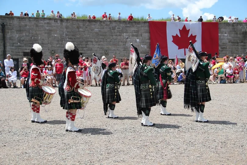 A line of bagpipers and drummers dressed in traditional Scottish attire, performing outdoors with a Canadian flag hanging in the background and an audience watching.