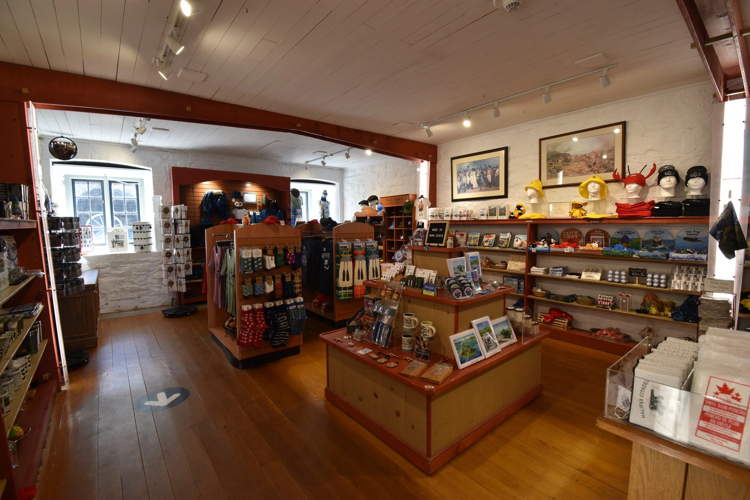 Interior of the Regimental Shop, a gift shop with wooden floors and white brick walls, displaying various souvenirs, clothing, and artwork.