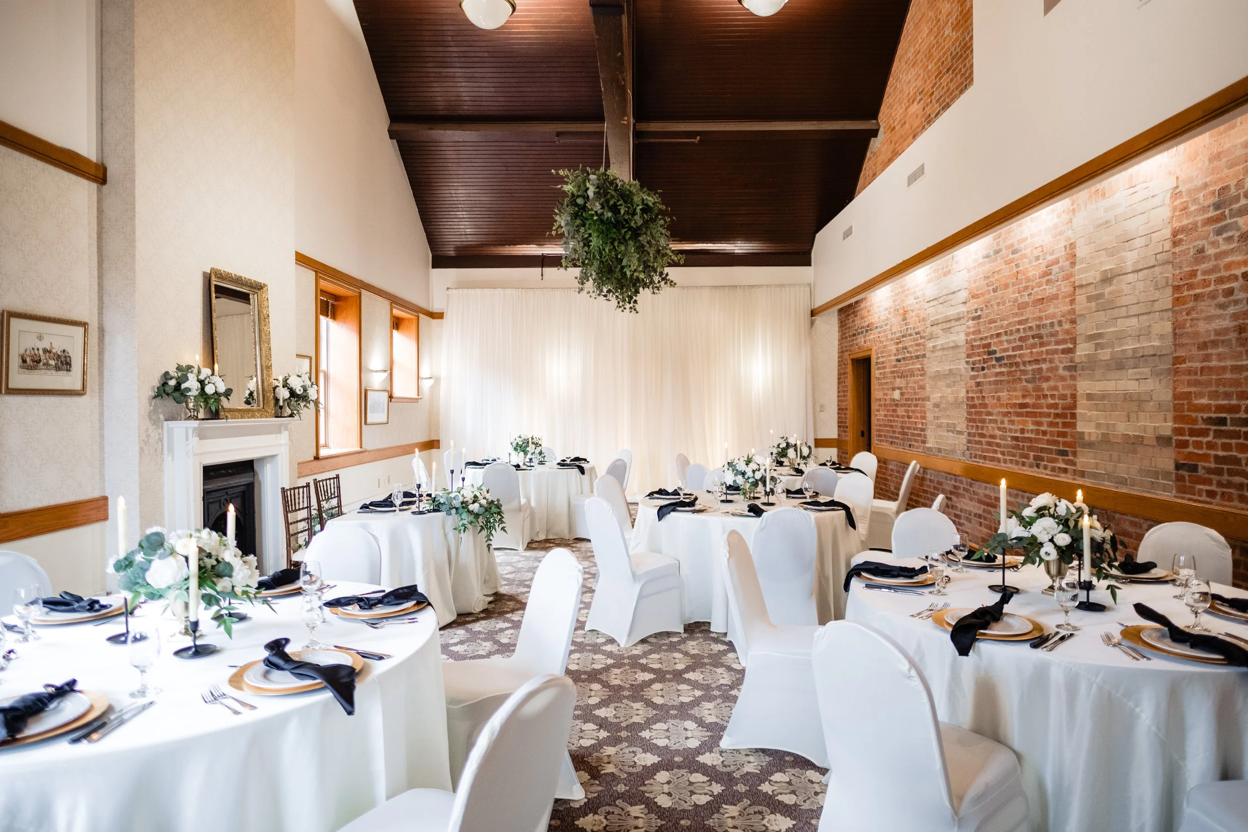 Elegant banquet hall decorated for a formal event with round tables covered in white tablecloths, each set with black napkins, candles, and floral centerpieces. The room features exposed brick walls, wooden accents, a high wooden ceiling, and a large greenery chandelier hanging from the ceiling.