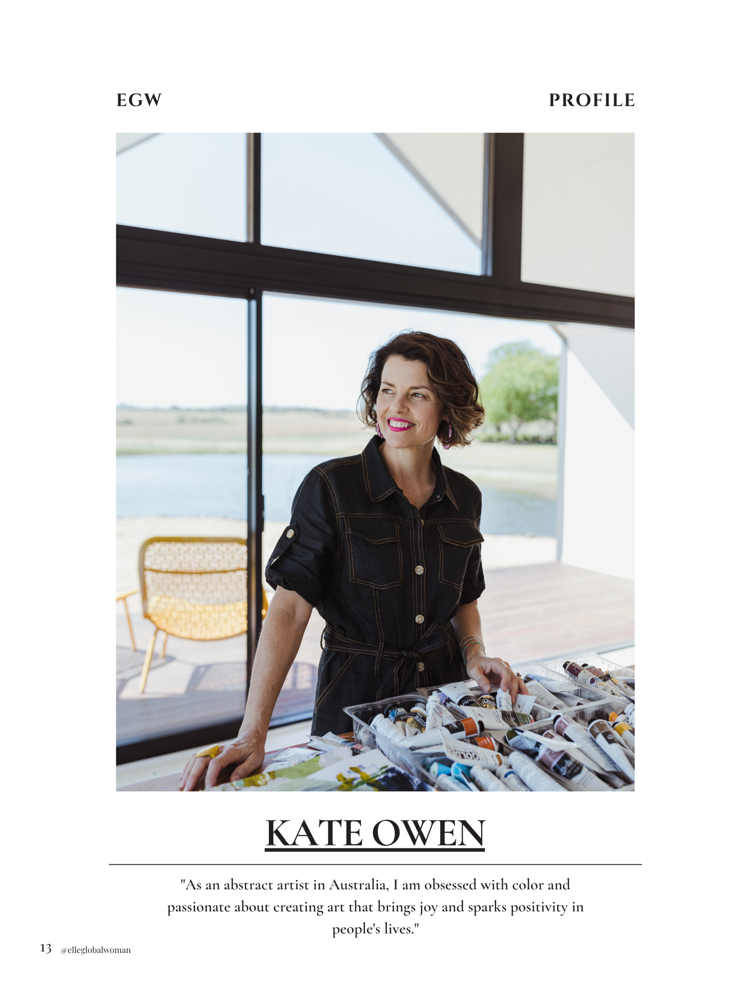 Photograph of a woman with curly brown hair, wearing a black shirt, standing behind a table with art supplies, in front of a large window with outdoor scenery in the background.