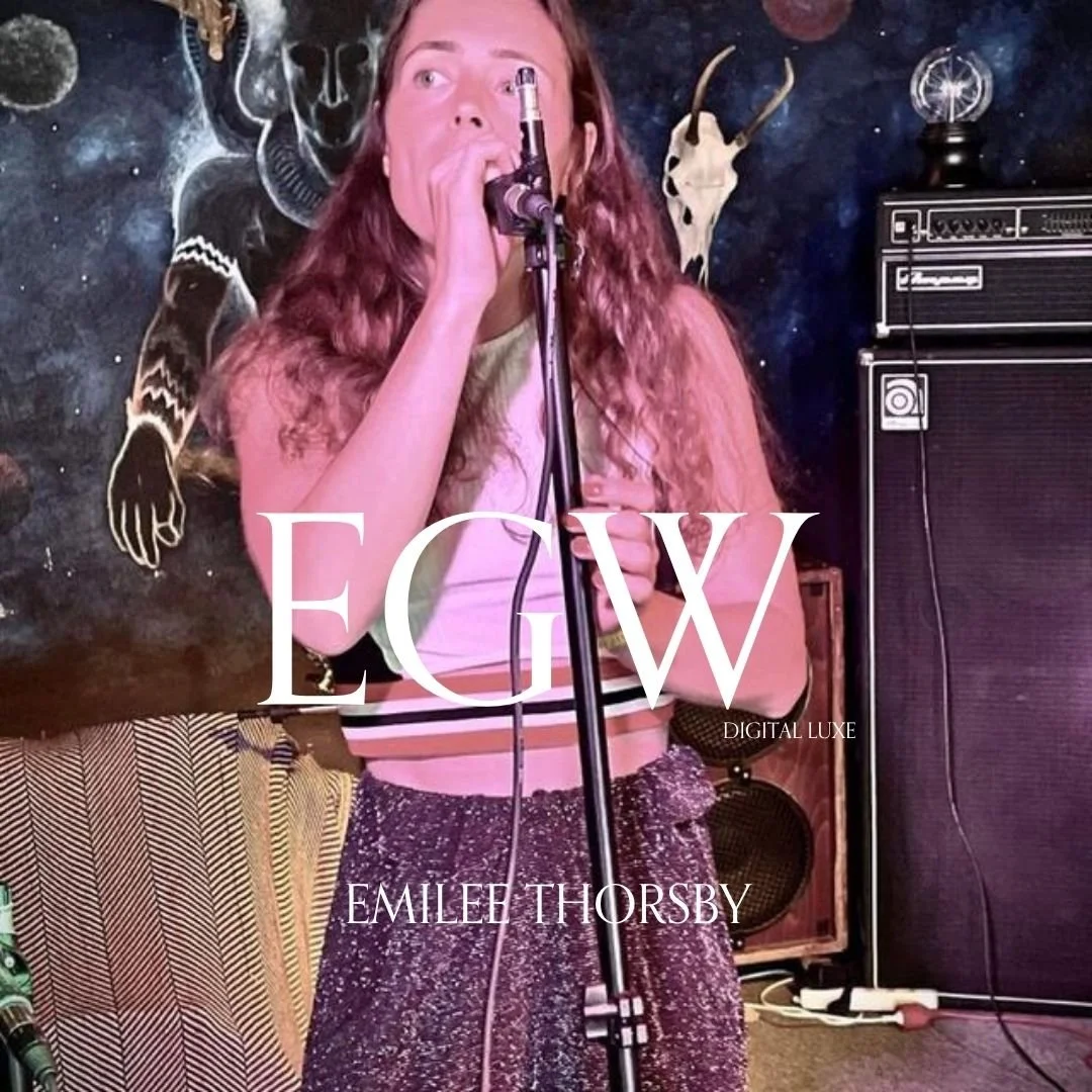 A woman with long curly hair singing into a microphone on stage, with a black background featuring star designs and a mounted animal skull with horns. She is wearing a pink crop top and high-waisted sparkling pants.