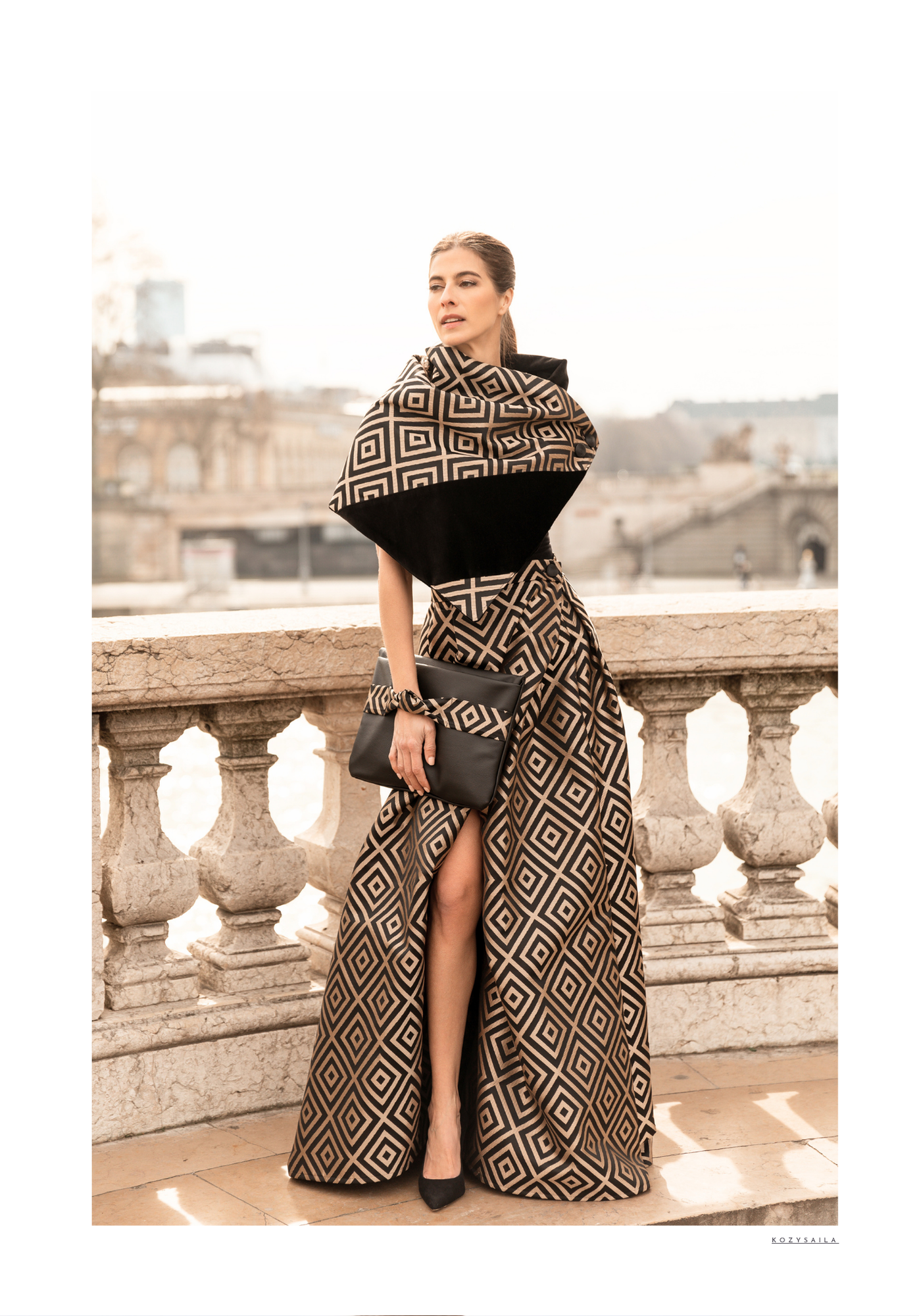 Woman standing on a stone balcony in a patterned dress and shawl, with a cityscape in the background.
