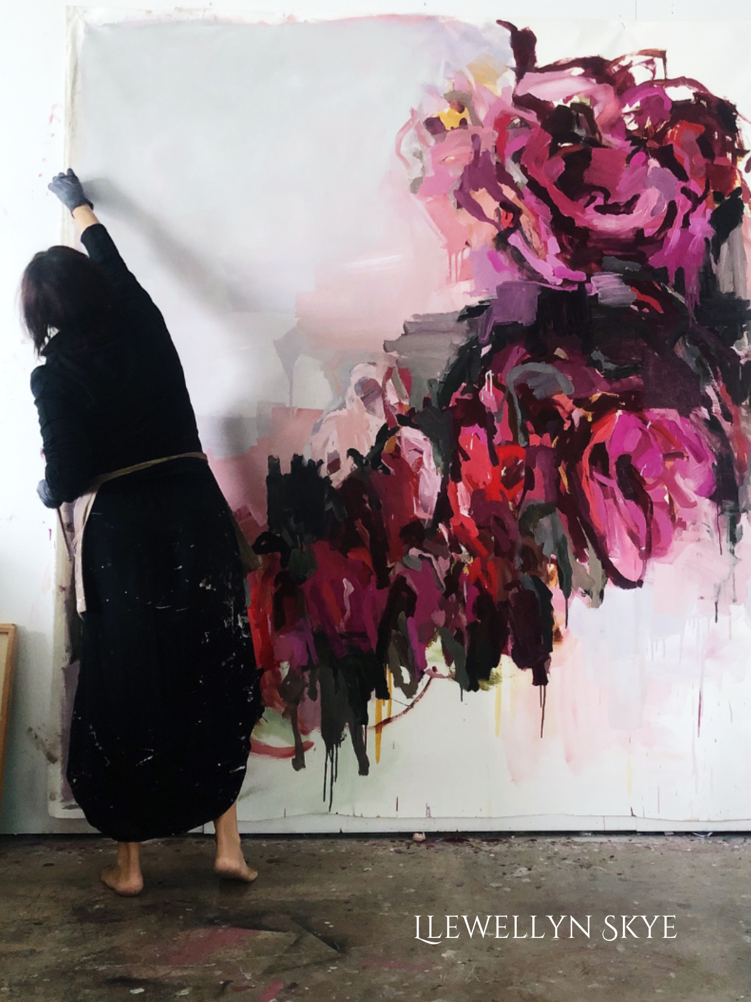 An artist working on a large, colorful floral painting of pink roses, dressed in black, barefoot, in an art studio with a concrete floor.