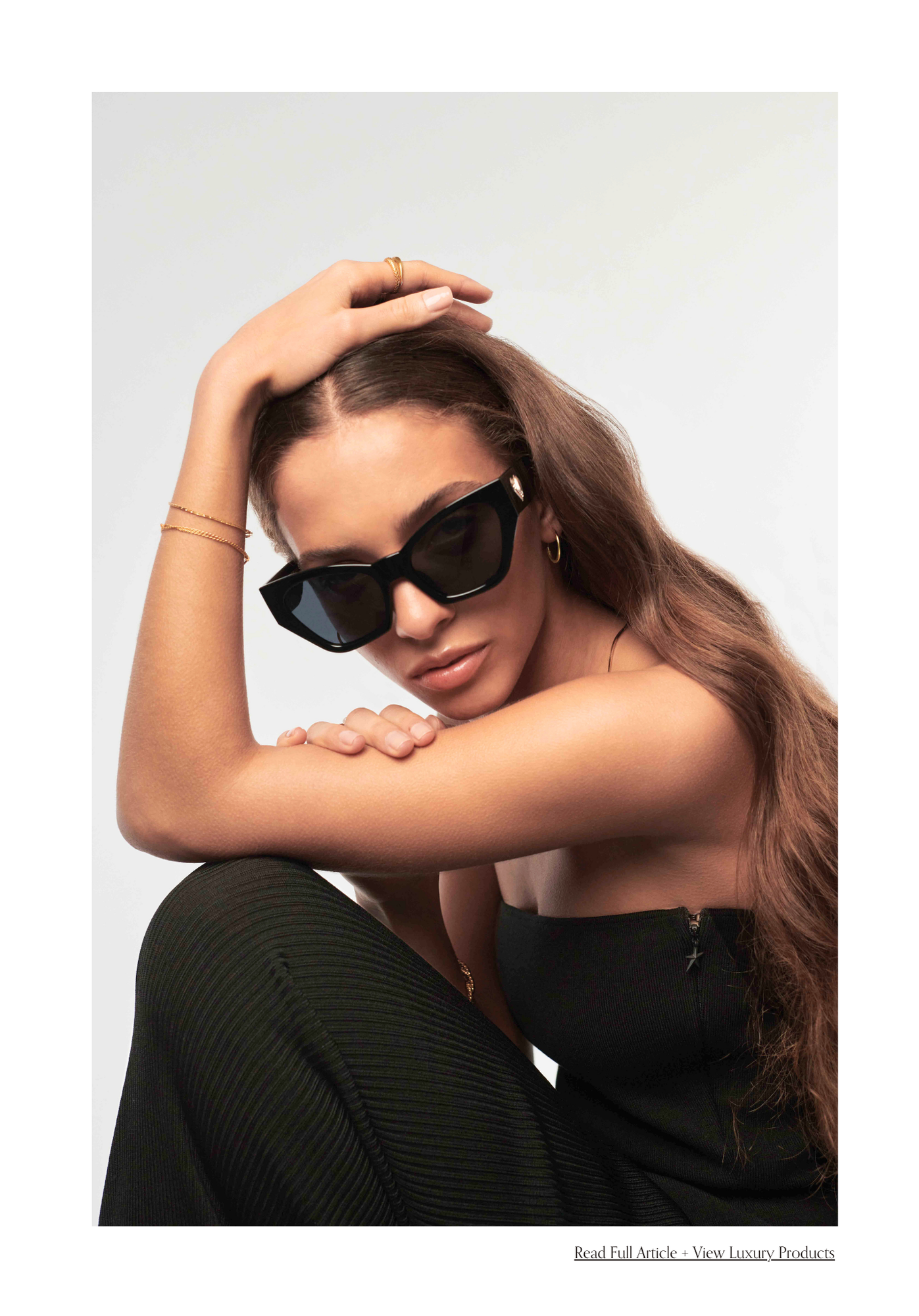 A woman with long brown hair and dark sunglasses, wearing a black strapless top, gold jewelry, and sitting against a plain white background.
