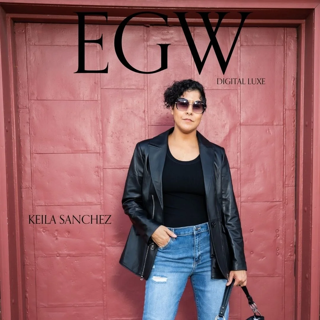 A woman standing in front of a pink wall, wearing sunglasses, a black leather jacket, a black top, and distressed blue jeans, holding a black handbag.