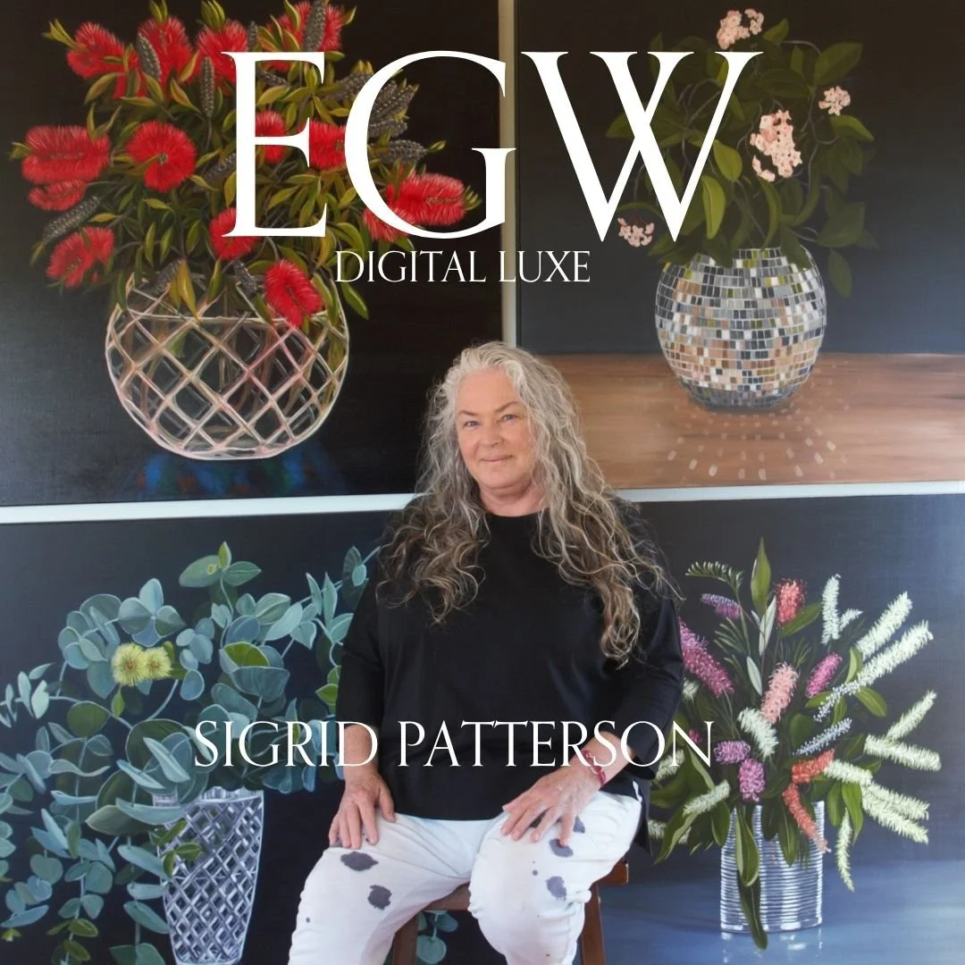 A woman with long, curly gray hair sitting in front of a background with large floral paintings and vases of flowers.