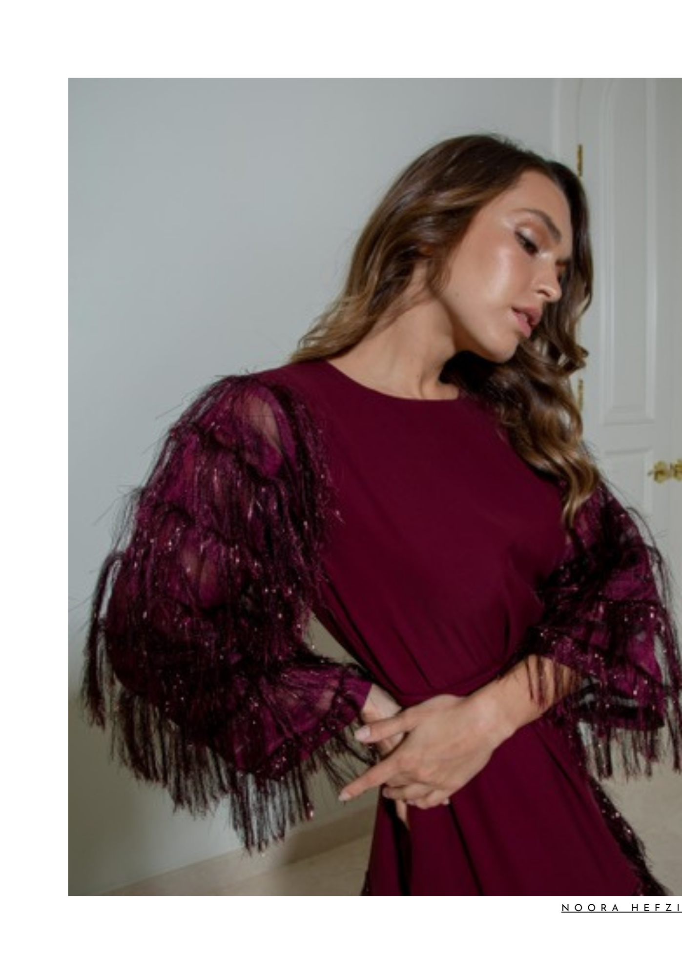 A woman with long brown wavy hair wearing a burgundy dress with lace and fringe details, standing with her eyes closed, arms crossed, in front of a white door.