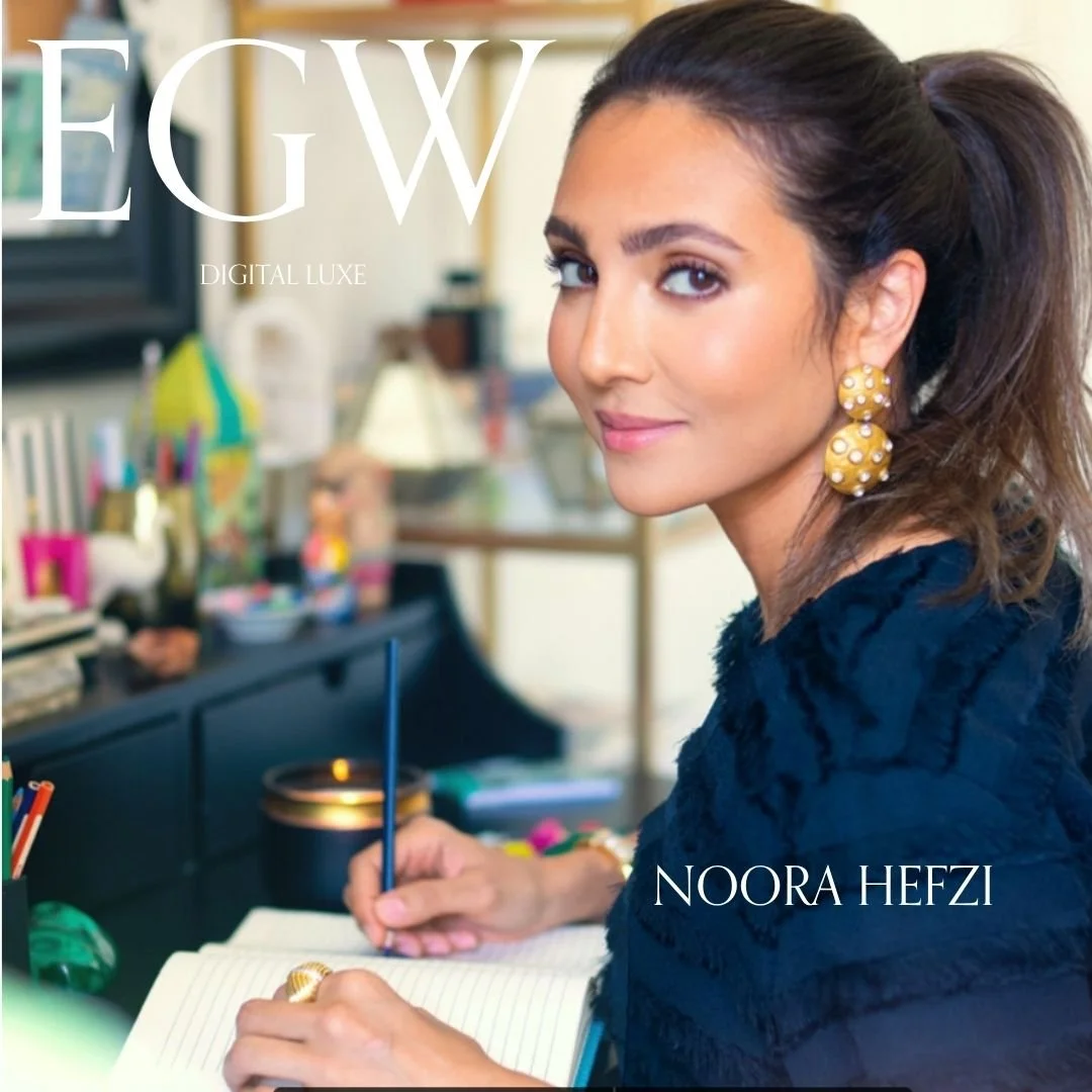 Cover image featuring a woman with brown hair tied back, wearing large yellow polka-dot earrings, sitting at a desk with a notebook and pen, smiling at the camera. The background shows a cluttered room with various colorful objects. The text on the i