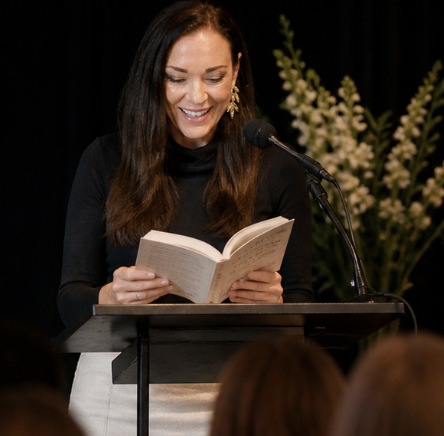 Woman smiling while reading a book at a podium with a microphone, in front of an audience and floral backdrop.
