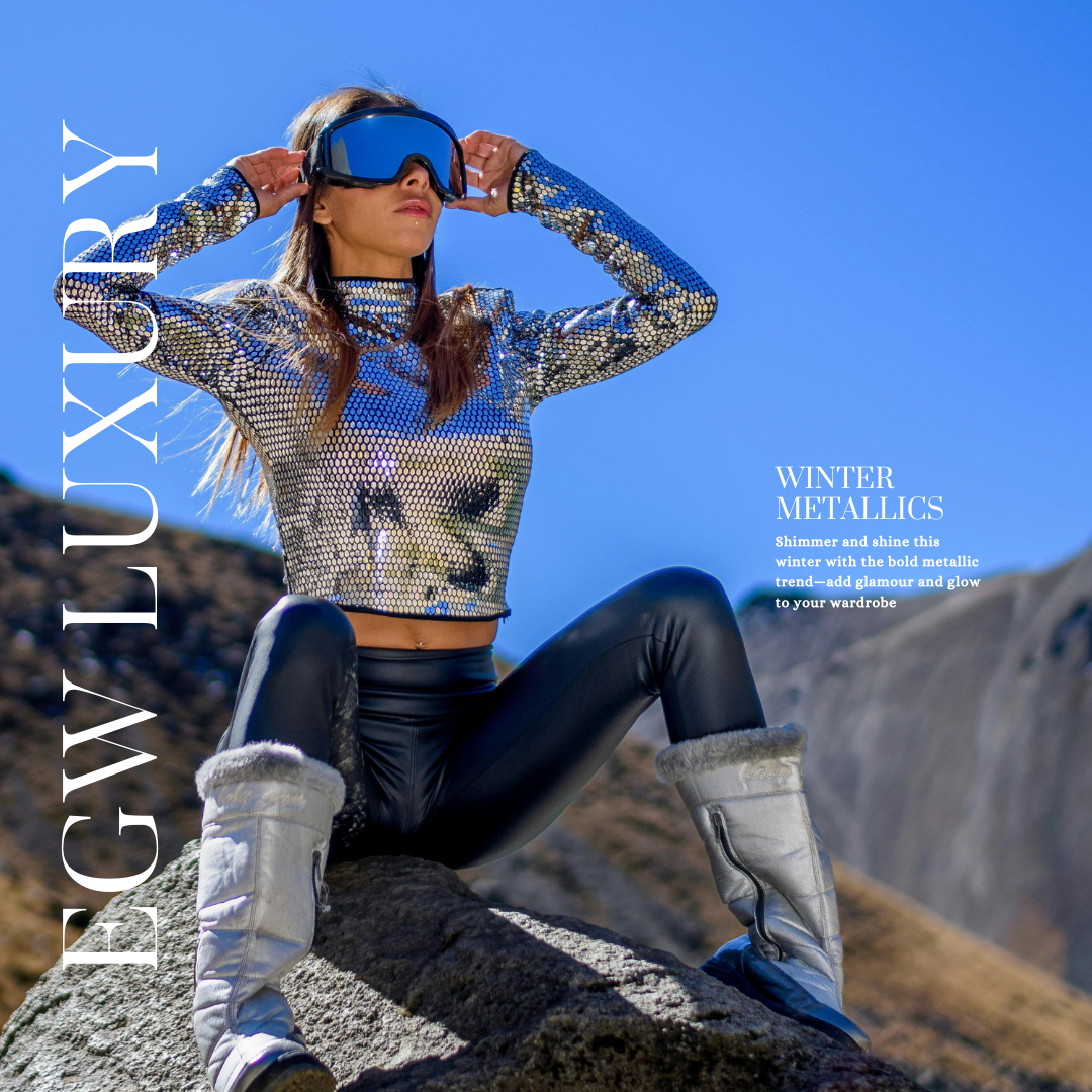A woman sitting on a rock outdoors wearing winter metallic clothing, including a shiny silver top, black leather pants, gray fur-lined boots, and ski goggles, with a clear blue sky in the background.