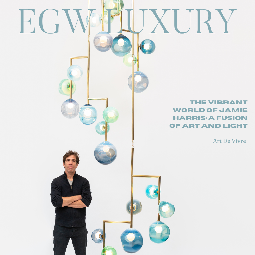 Cover of a magazine titled "EGW LUXURY" featuring a man standing with arms crossed in front of a white background. Above him is a modern art installation with hanging glass orbs in various shades of blue, green, and clear, suspended by gold rods. The