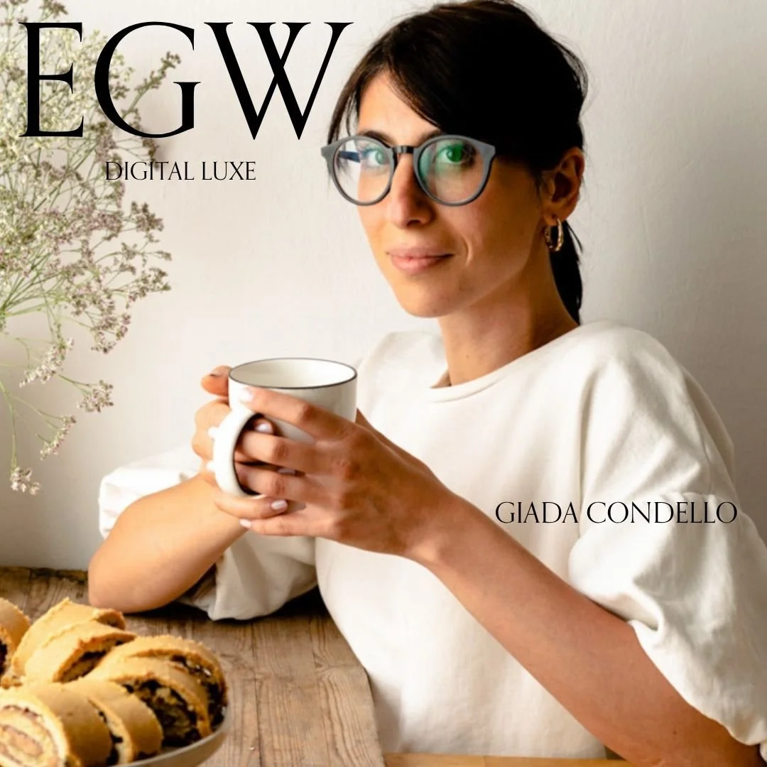 A woman with dark hair and glasses holding a white mug, sitting at a wooden table with a plate of rolled cookies, and a vase with flowers in the background, accompanied by text that includes 'EGW' and 'GIADA CONDELLO'.