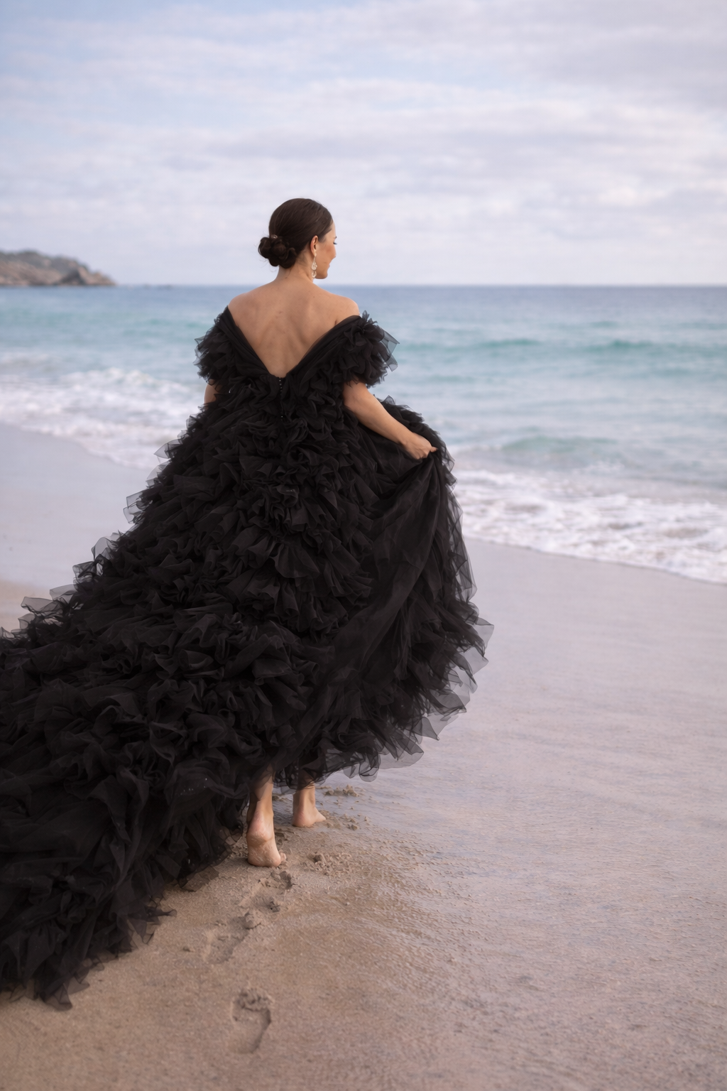 Woman in a black ruffled gown walking barefoot on the beach near the ocean with a cloudy sky overhead.