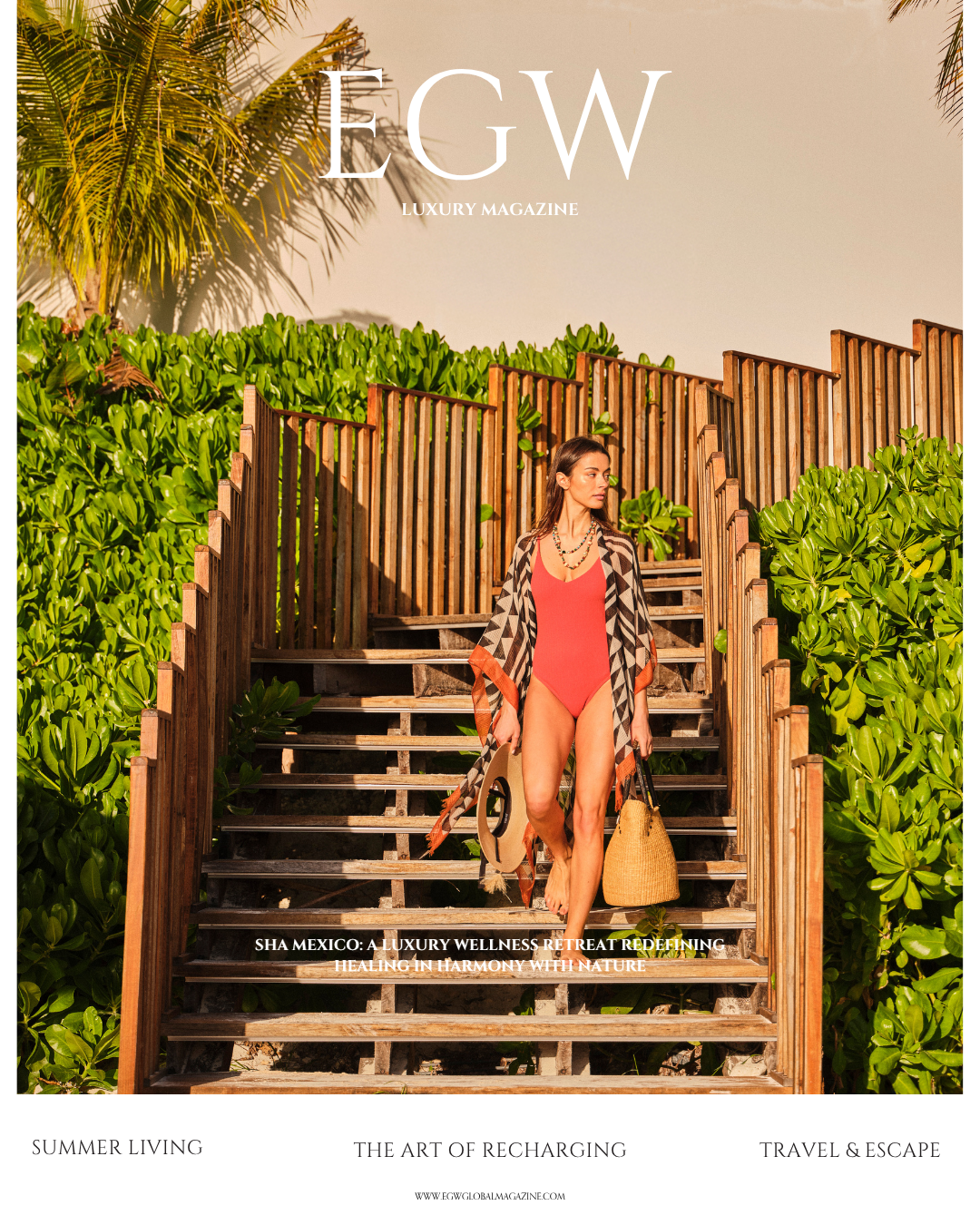 A woman in a coral swimsuit and a patterned cover-up walking down wooden stairs on a tropical beach, carrying a large straw beach bag and a hat, with lush green bushes and palm trees in the background.