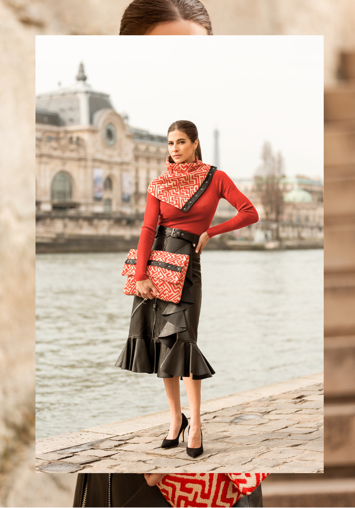 A woman standing near the water's edge on a stone walkway, wearing a red long-sleeve top, black ruffled leather skirt, high heels, and holding a patterned clutch purse with a matching scarf. An ornate building with a clock tower is visible in the bac