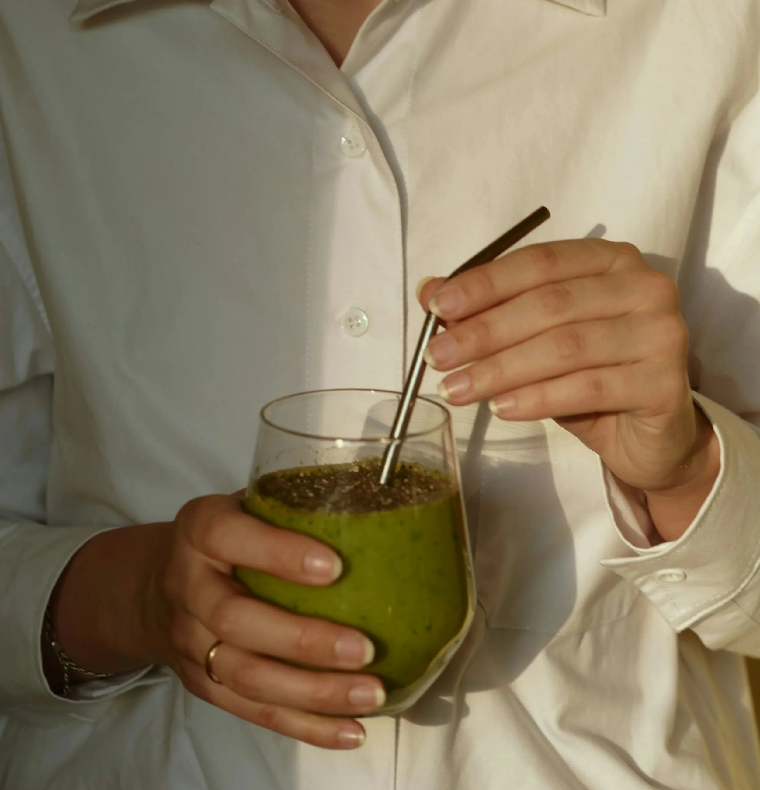Person wearing a white shirt holding a glass of green smoothie with a metal straw.