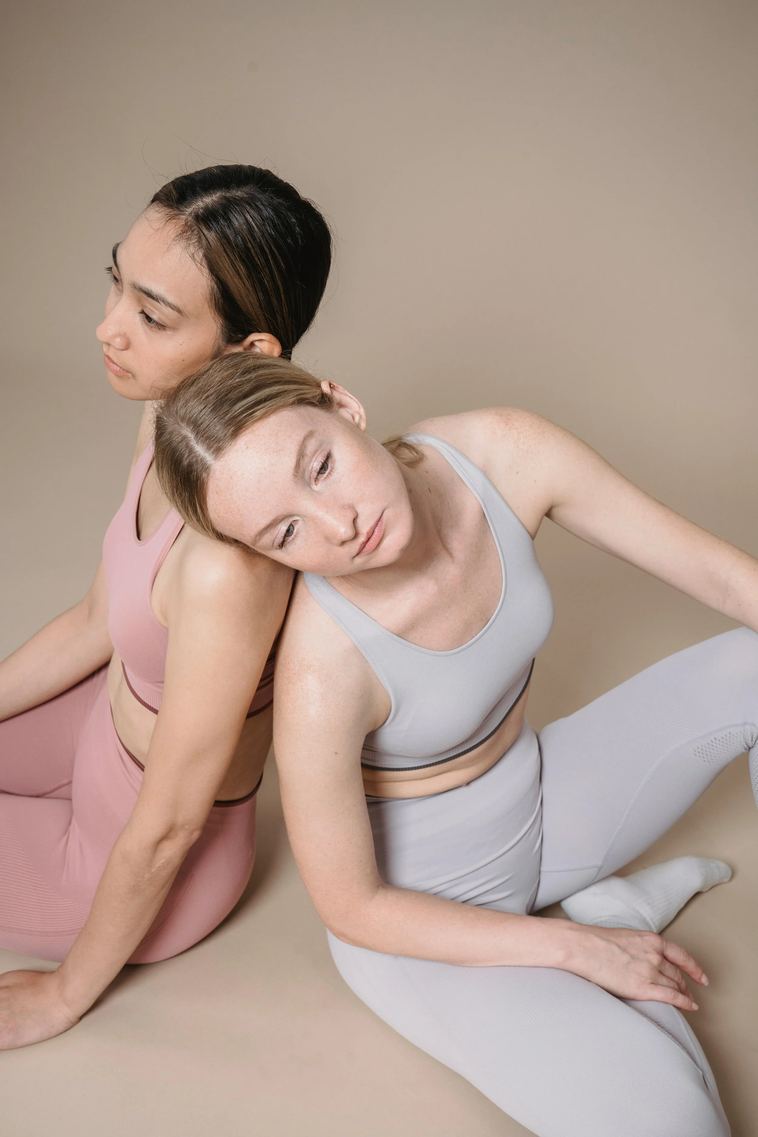 Two women in pastel-colored workout clothes sitting back-to-back on a beige floor with neutral background, one resting her head on the other's shoulder.