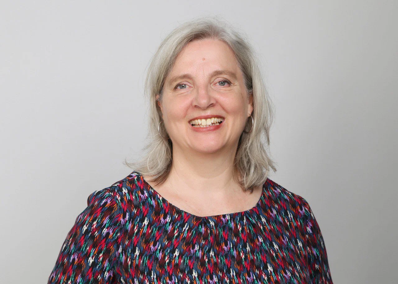 Lächelnde ältere Frau mit grauem Haar und buntem Oberteil vor weißem Hintergrund.