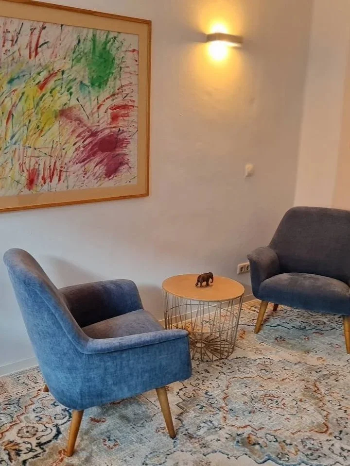 Ein Wohnzimmer mit zwei dunkelblauen Sesseln, einem kleinen runden Tisch mit einer Elefantenfigur, einem bunten abstrakten Gemälde an der Wand und einem Wandlicht. Es steht ein Teppich auf dem Boden.