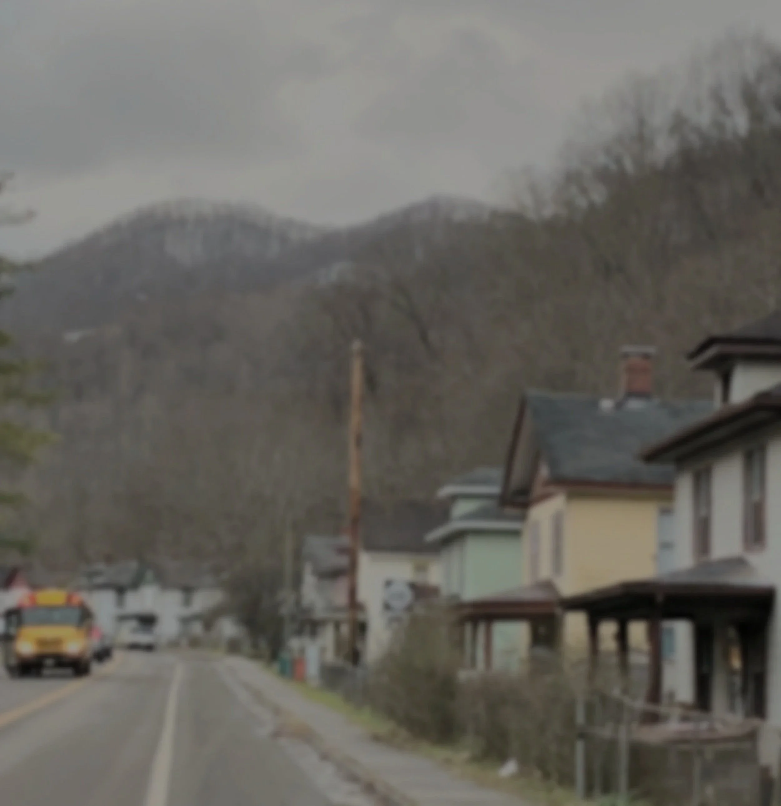 A blurry image of a street scene in an Appalachian town showing parked cars, buildings, and trees, taken during a cloudy day.