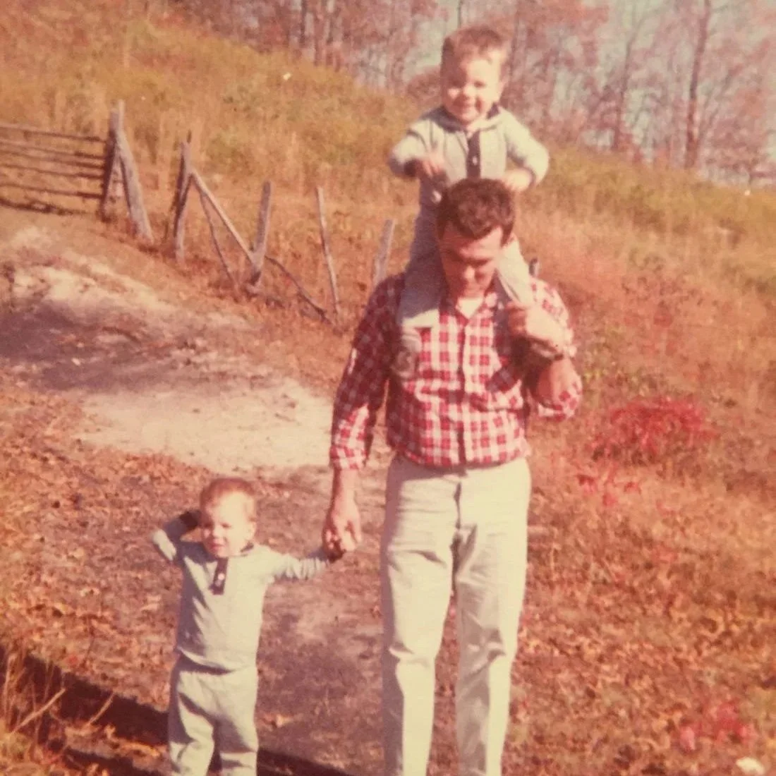 A man carrying a young boy on his shoulders, holding hands with a small child while walking on a dirt path surrounded by autumn leaves.