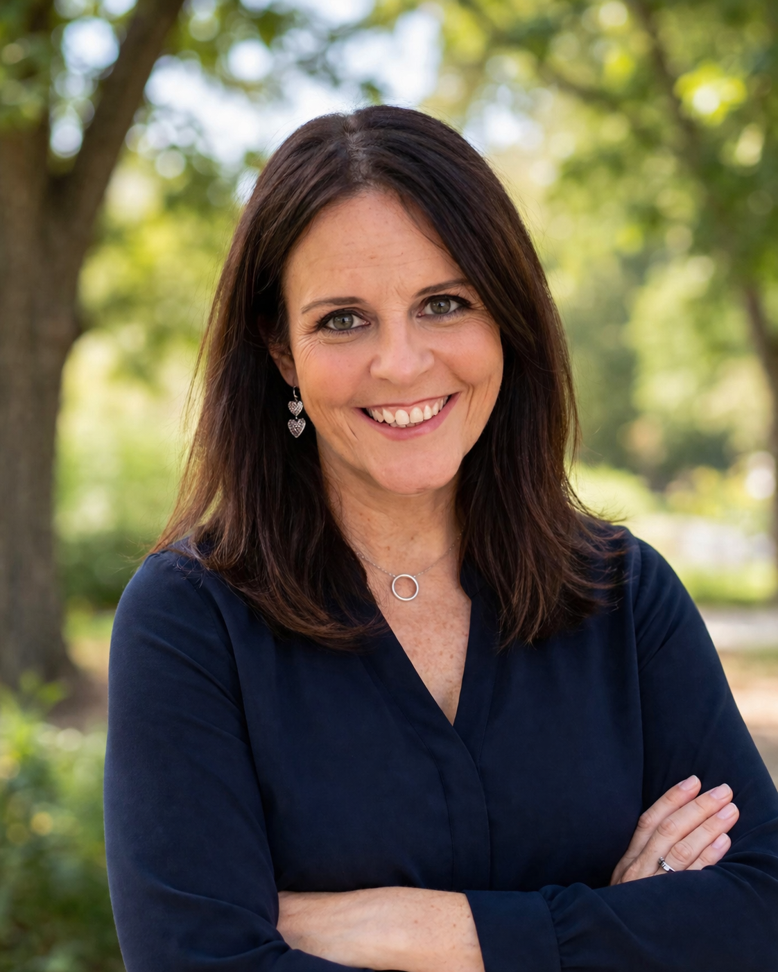 A woman with brown hair and blue eyes smiling outdoors, wearing earrings and a dark blue blouse, with trees and sunlight in the background.