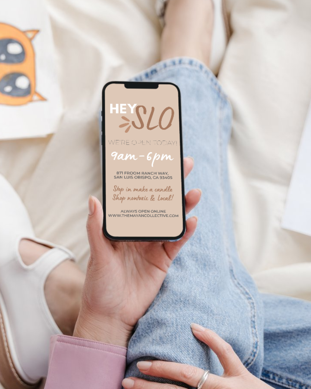 A person holding a smartphone displaying an opening hours sign for 'HEY SLO' with the text 'We're open today! 9am-6pm,' an address in San Luis Obispo, CA, and a note about online shopping.