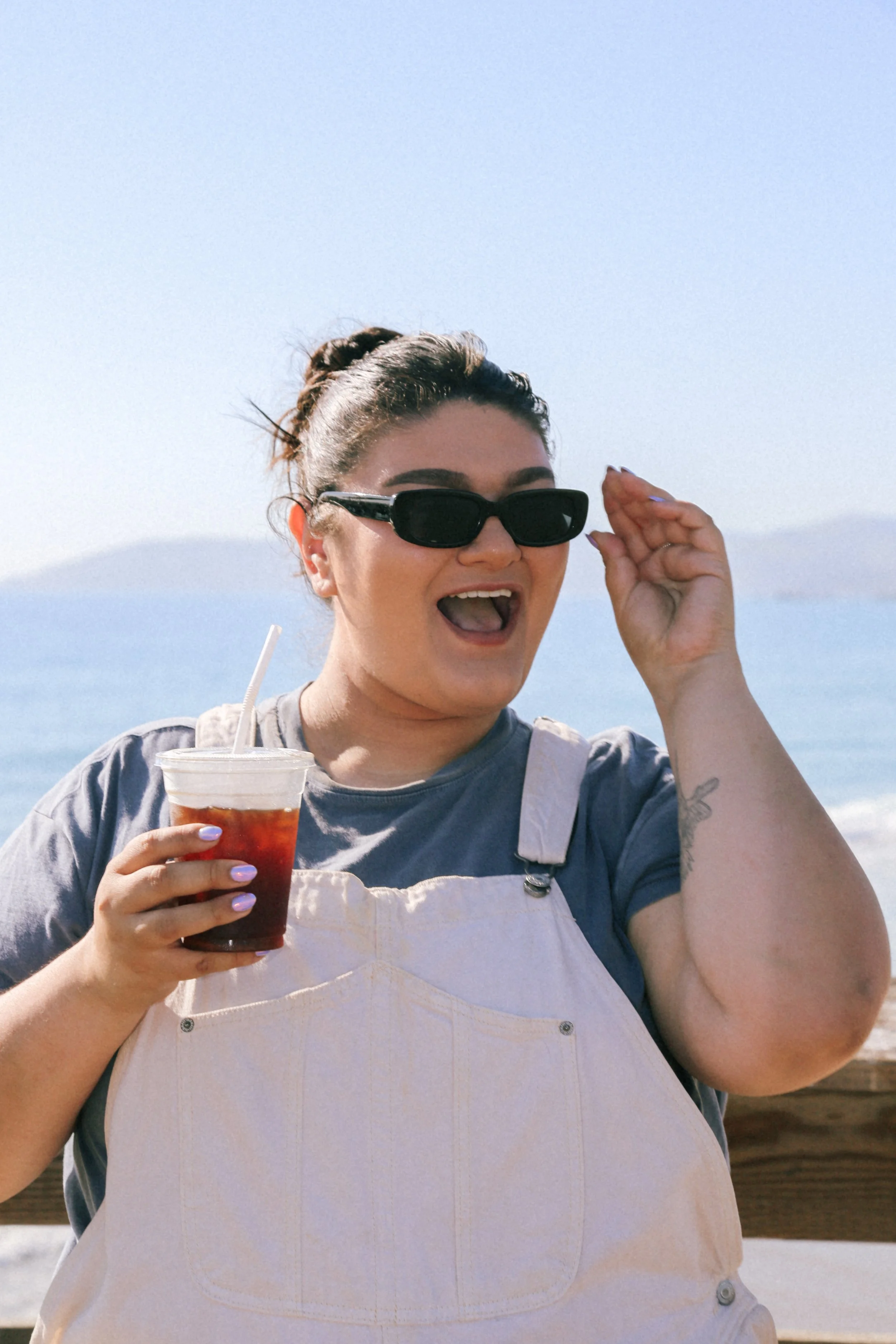 A woman with dark hair in a bun wearing sunglasses, a gray t-shirt, and cream overalls is smiling by the beach with an ocean in the background, holding a cold drink.