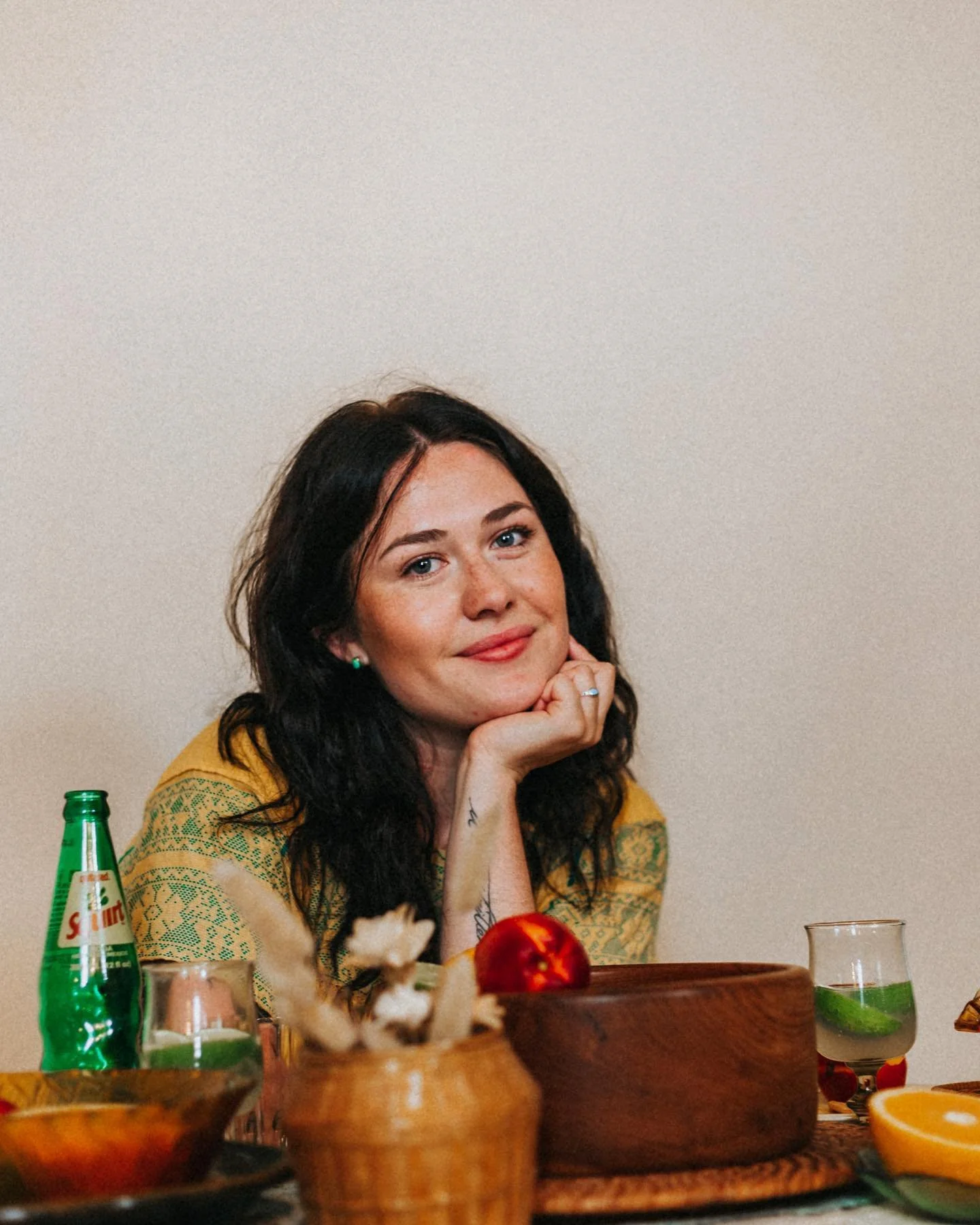 A woman with black hair and blue eyes, wearing a yellow-patterned top, sitting at a table with food and drinks in front of her, resting her chin on her hand, smiling, against a plain beige background.