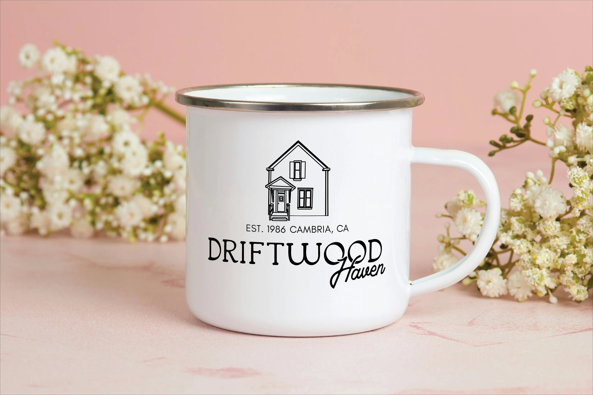 A white mug with black outline of a house and text that reads "Driftwood Haven." The mug is surrounded by white flowers with a pink background.