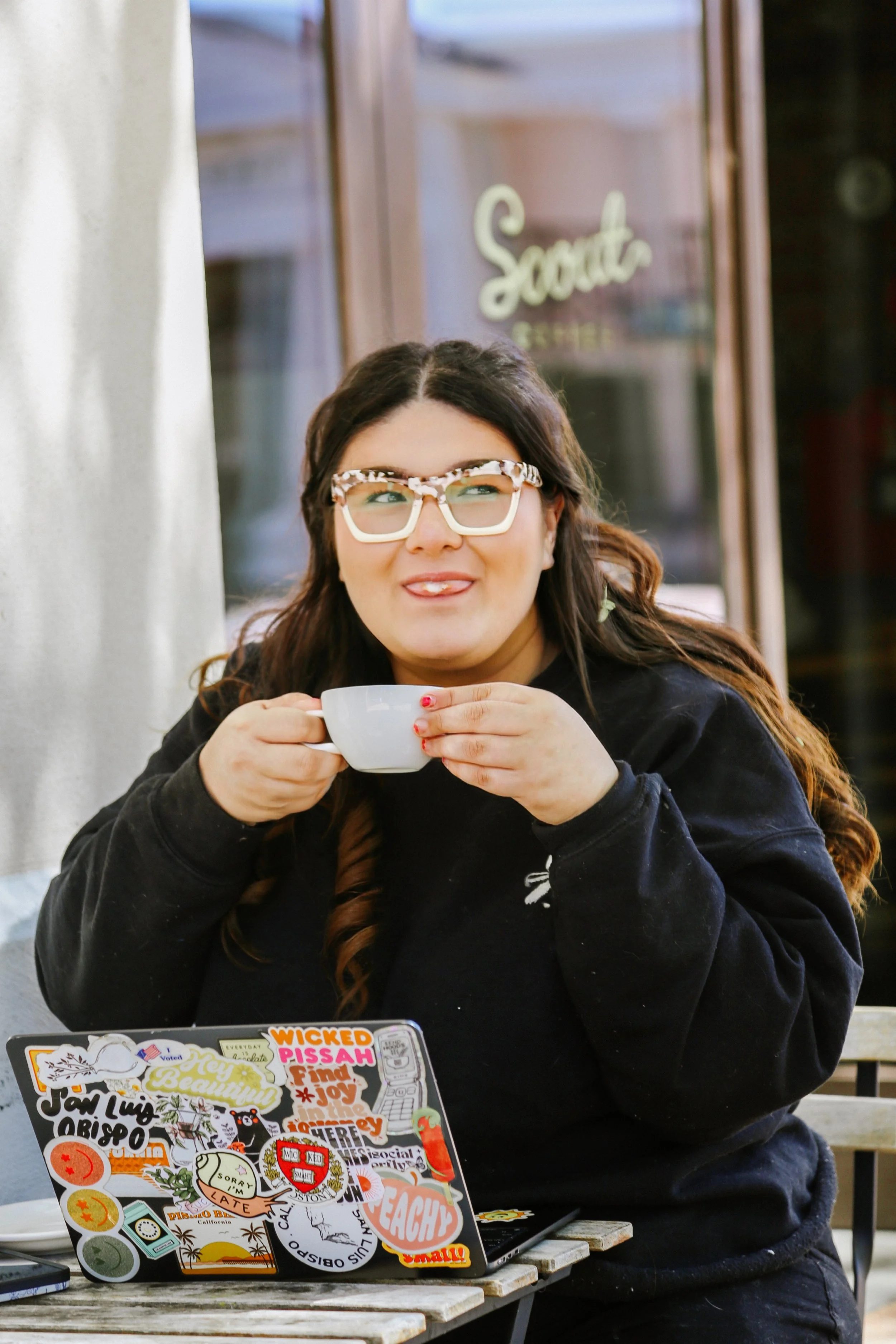 A woman with long dark hair and glasses with a patterned frame sitting at a table, holding a white mug, with a laptop covered in colorful stickers in front of her, on an outdoor patio.