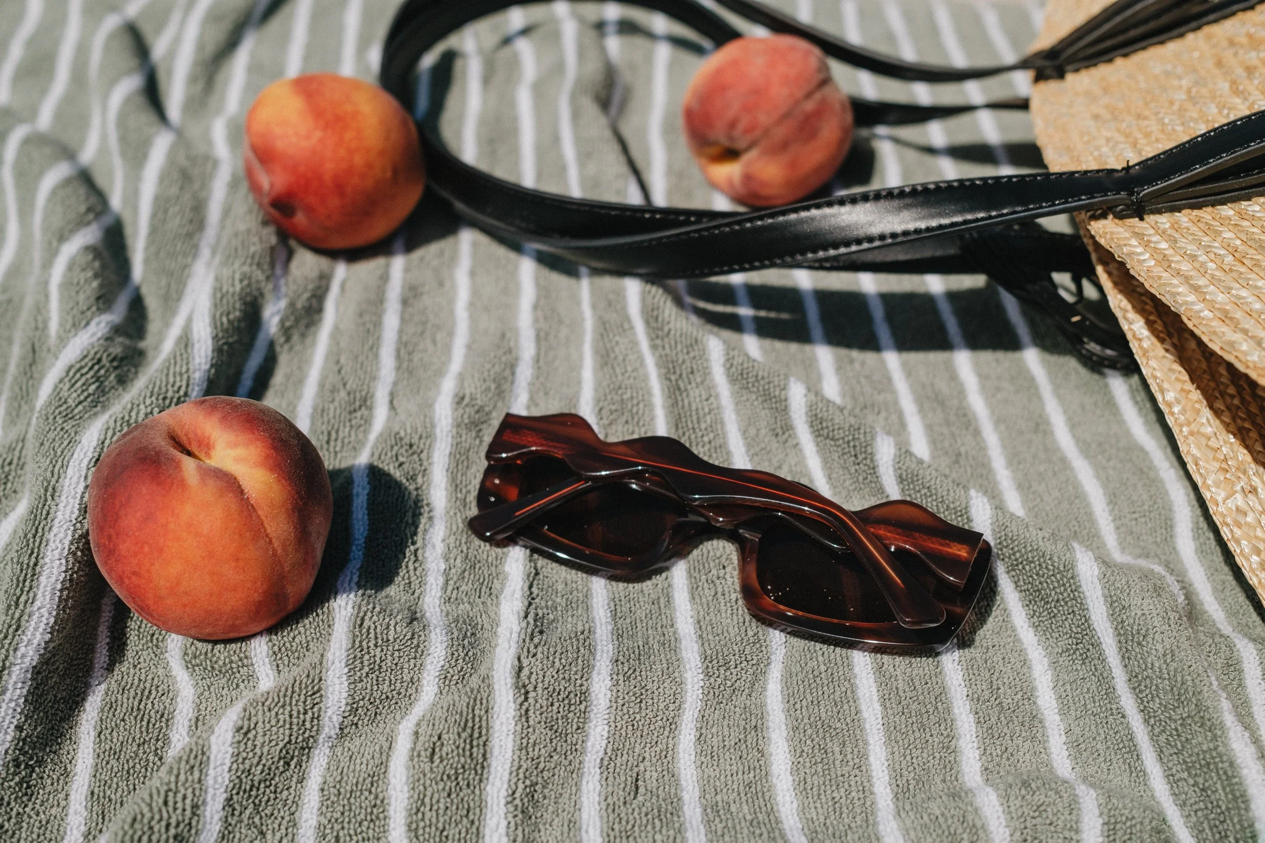 Three peaches, a black scrunchie, a brown hair clip, and parts of a straw bag on a striped green and white cloth.