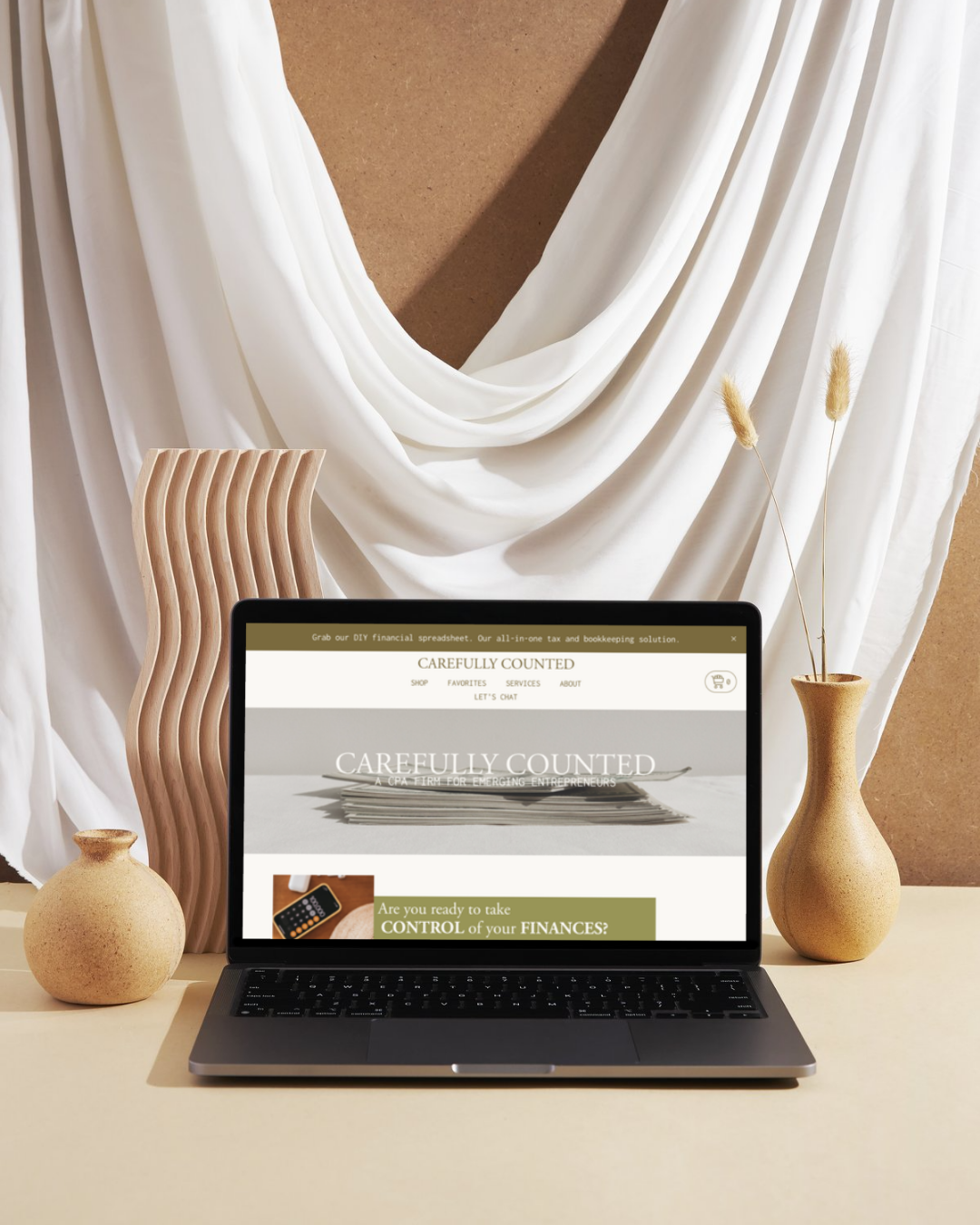 Open laptop on a light-colored desk displaying a financial website, flanked by two beige ceramic vases, with stone and wooden accents, white draped fabric backdrop.