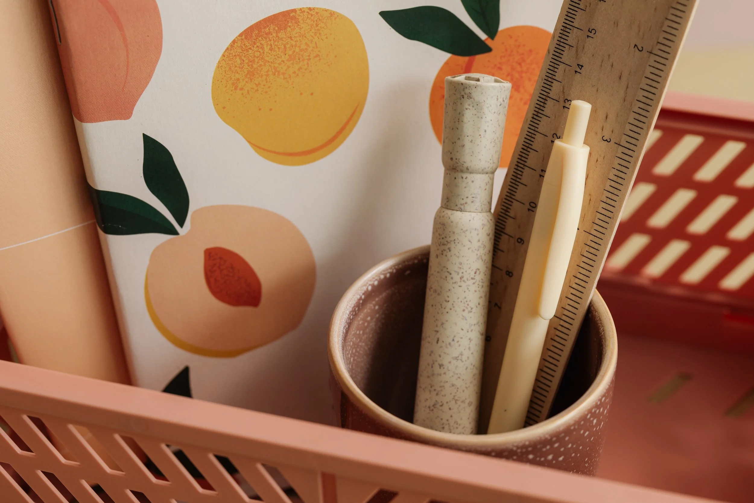 A pink storage basket with a cup holding a speckled pen, a beige pen, and a ruler, with a wall decorated with abstract fruit illustrations in the background.